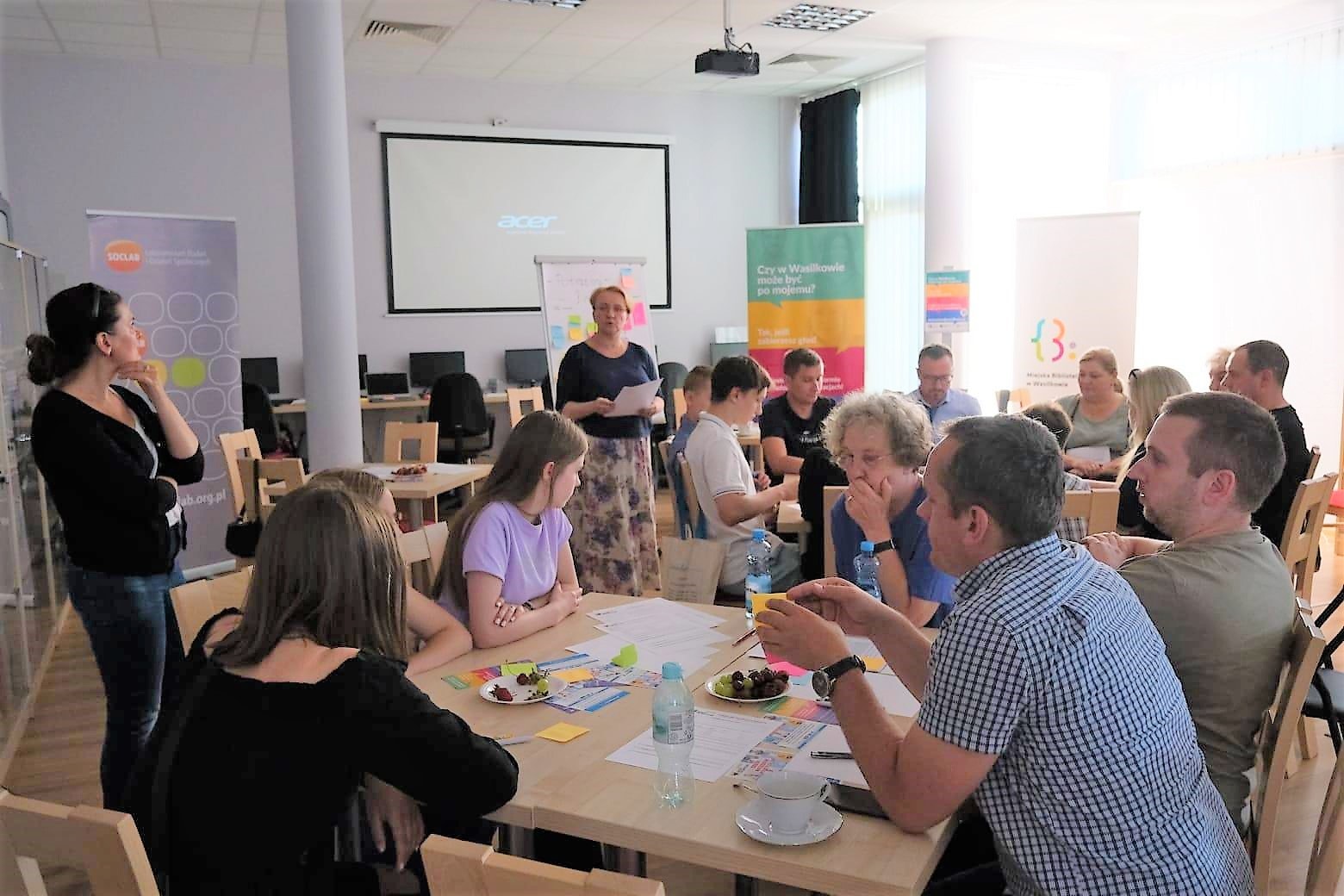 Kilkanaście osób na warsztatach, siedzą przy stolikach.